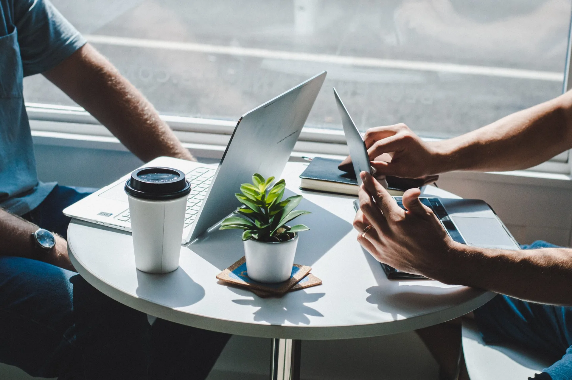 Two colleagues working across a small table with laptops in a sunlit office